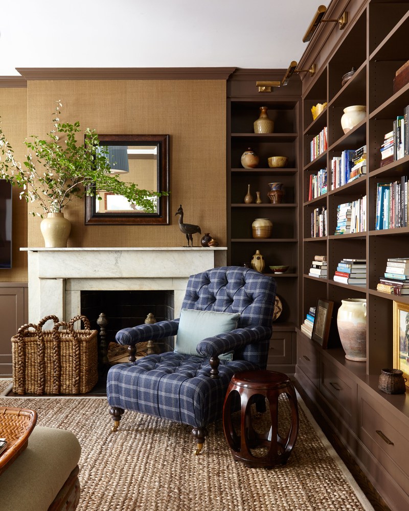 Library with navy blue plaid armchair, marble fireplace, firewood basket, jute rug, brown built-in bookcases
